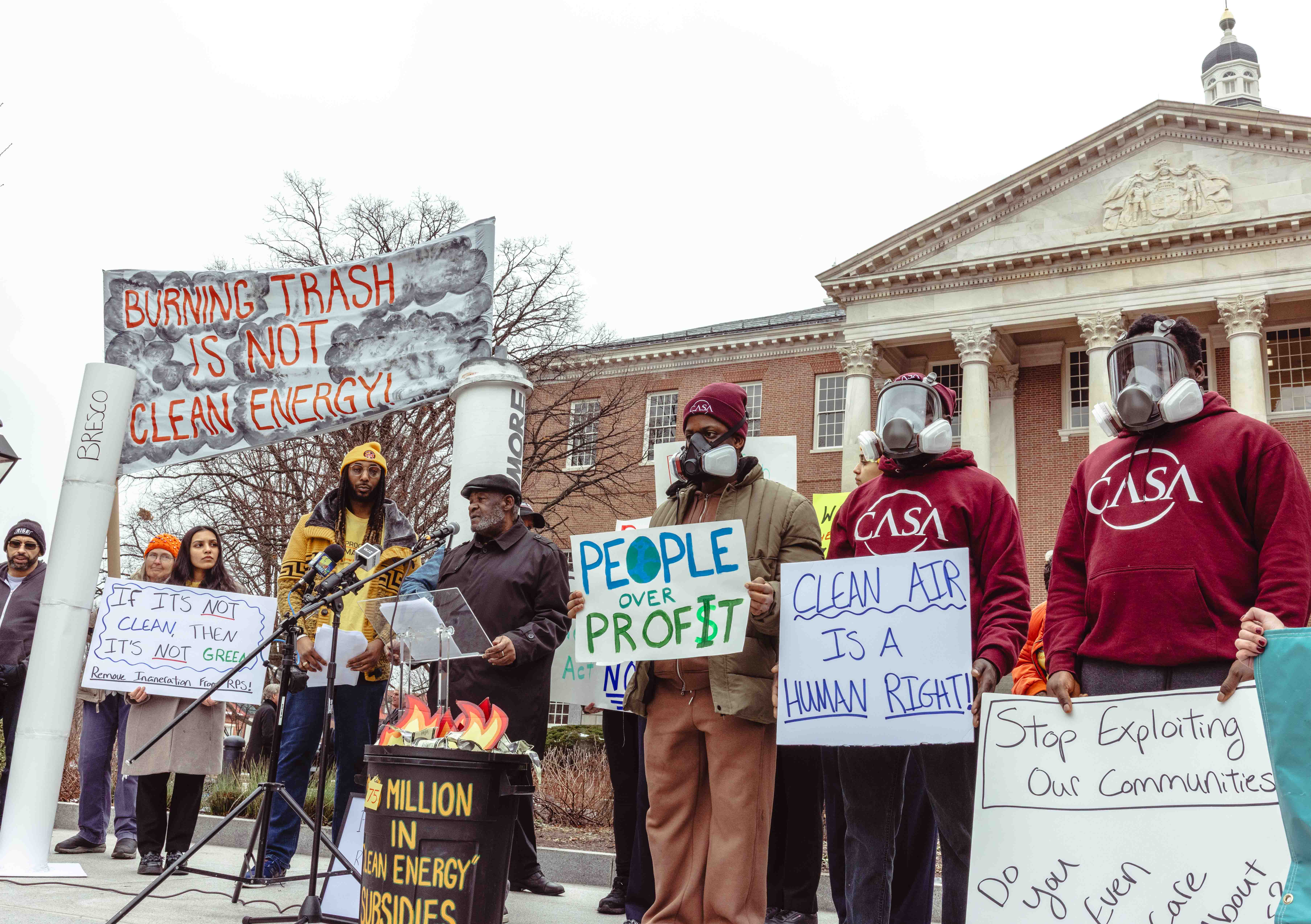 People rallying in front of the MD State House, some wearing gas masks, with signs like "Burning trash is not clean energy," "clean air is a human right," and "people over profit."