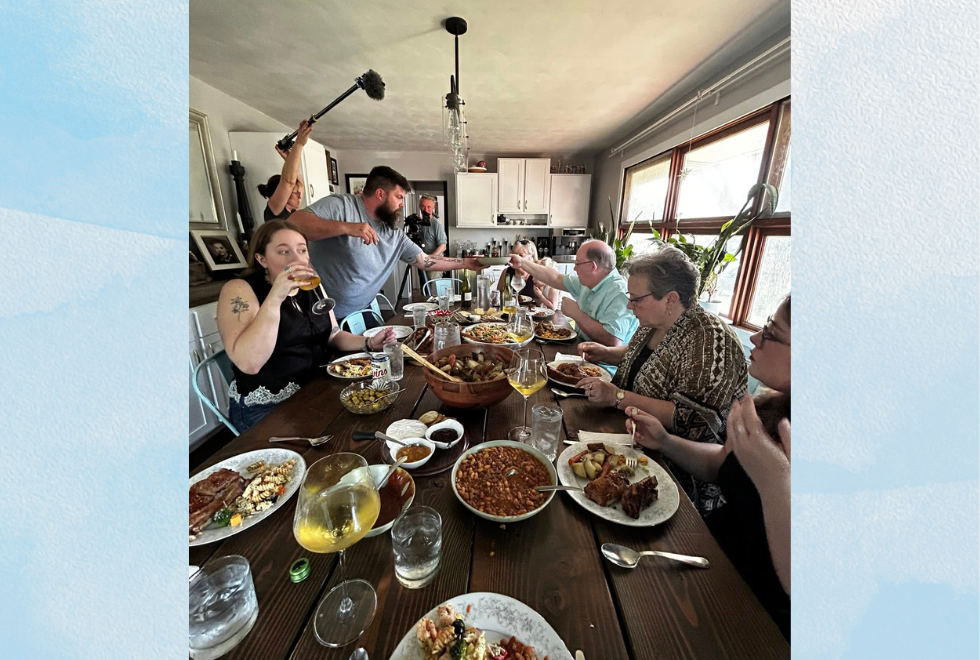 A dozen people sit for dinner at a home, with a small film crew setting up equipment around the room.