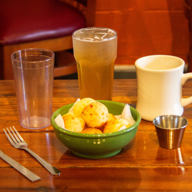 Ceramic bowl with pao de queijo surrounded by reusable cups, silverware, and sauce cups