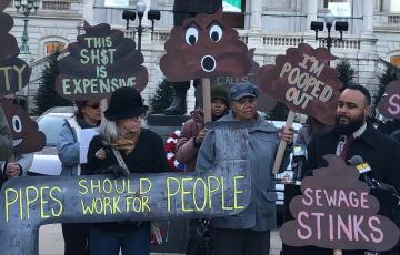 A crowd of people rallying outside of Baltimore City Hall, with signs like "Pipes should work for people" and "sewage stinks"