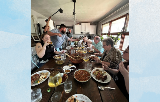 A dozen people sit for dinner at a home, with a small film crew setting up equipment around the room.